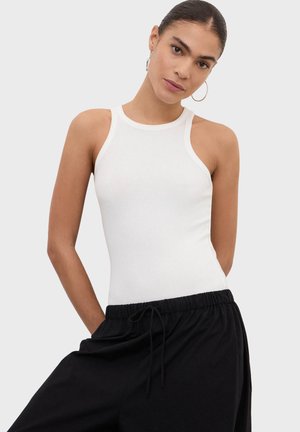 Žena s vlasmi stiahnutými dozadu, ktorá nosí kruhové náušnice, biele bezrukávové tričko a čierne nohavice sťahované šnúrkou, pózuje s rukami v vreckách.