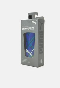 Guardia di calcio PUMA in un'imballaggio grigio. Presenta un motivo geometrico blu e verde con una finestra trasparente e un gancio per appenderlo. Taglia L.