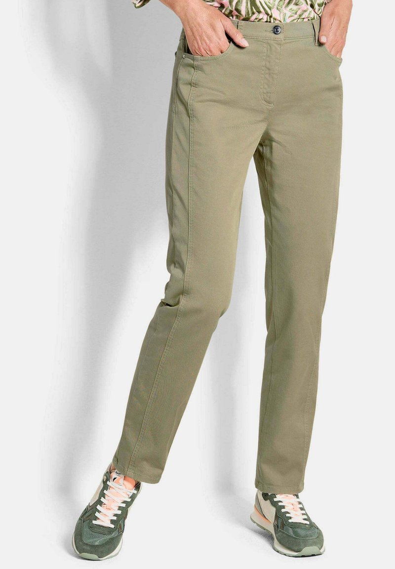 Person trägt beige gerade geschnittene Hosen und grüne Sneakers mit weißen Schnürsenkeln, die Hände ruhen in den vorderen Taschen.