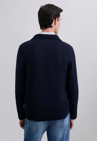Cárdigan de punto azul marino con cuello chal, puños acanalados y dobladillo. Lleva puesto sobre una camisa de color claro y combinado con jeans azules.