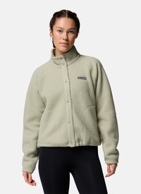 Young woman with braided hair wearing a light green button-up fleece jacket and black leggings, facing forward against a plain background.