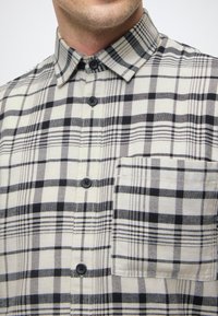 Koszula w kratę w odcieniu kremowym z czarnymi i szarymi wzorami, z zapięciem na guziki, z kołnierzykiem o spiczastym kształcie oraz pojedynczą kieszonką na piersi.