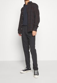 Zwart-oranje geruit overhemd met knopen over een marineblauwe trui, gecombineerd met grijze broek met patroon en zwarte sneakers met witte zolen.