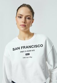 Witte hoodie met zwarte tekst, waarop "San Francisco," "Sporty 75 Golden Gate" en "North West College" staat. Ronde halslijn en een losse pasvorm.