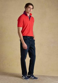 Jeune homme debout, une main dans la poche, portant un polo rouge à col relevé, un jean foncé et des baskets bleu marine.