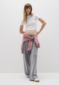 Weißes, gekürztes T-Shirt, graue weit geschnittene Hose mit Nahtdetails und ein pinker Pullover, der um die Taille gebunden ist. Heller Hintergrund, lässiges Outfit.