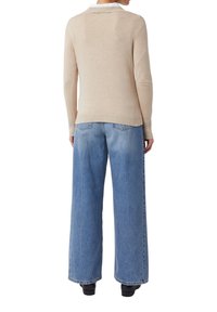 Person, der einen beigen Pullover und weite blaue Jeans trägt, steht mit dem Rücken zur Kamera vor einem weißen Hintergrund.