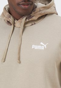Sudadera beige con capucha de patrón camuflaje, con un logo blanco de Puma en el pecho y cordones. Tejido suave con un acabado texturizado.