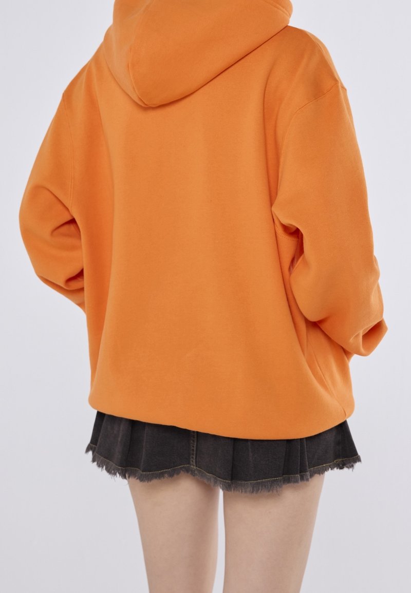 Felpa arancione oversize con cappuccio, realizzata in tessuto morbido. Abbinata a una mini gonna pieghettata nera, con dettagli di orlo sfrangiato.