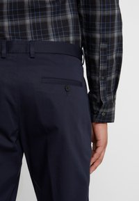 Mörkblå byxor med en jämn textur, med en sidoficka som är stängd med en knapp, i kombination med en svart och blå rutig skjorta.
