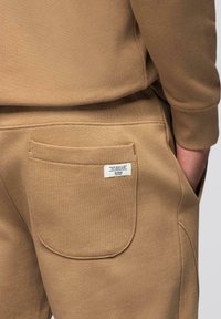 Poche arrière d'un pantalon de survêtement marron avec une petite étiquette blanche indiquant "VOTRE SEUL LIMITE EST VOTRE ESPRIT" et une partie de la main d'une personne dans la poche.