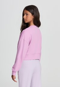 Felpa cropped in pile rosa chiaro, collo rotondo, spalle scese e polsini a coste, abbinata a pantaloni coordinati viola chiaro.