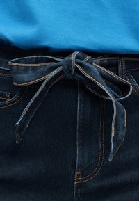 Ζώνη από τζιν δεμένη με φιόγκο γύρω από ένα ζευγάρι σκούρα μπλε τζιν με πορτοκαλί ραφές, φορεμένη με ένα φωτεινό μπλε πουκάμισο που είναι μπουστάκι μέσα στη μέση.