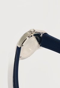 Ασημένια θήκη ρολογιού με χαραγμένη πίσω όψη, μπλε λουρί από υφή καουτσούκ και κορόνα στο πλάι, εμφανίζεται σε λευκό φόντο.