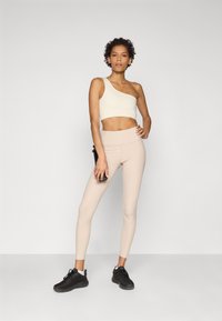 Gaiši bež viena pleca crop top, apvienots ar kailu ribotu legingu un melniem sporta apaviem. Apģērbs izceļas ar elegantu dizainu un pieguļošu siluetu.