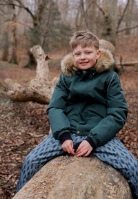 A child sits on a log wearing a dark green padded jacket with a fur-lined hood and patterned blue ski pants, hands resting on the log.