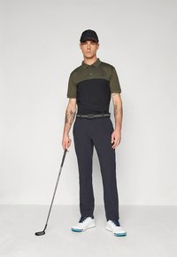 Golfoutfit med en svart och olivgrön pikétröja, mörka byxor, ett bälte och vita skor med blå detaljer. Modellen håller i en golfklubba.