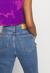 Jeans de mezclilla azul de cintura alta con un lavado claro, que cuentan con cinco bolsillos y una etiqueta de cuero color marrón. La tela tiene una textura suave y resistente.
