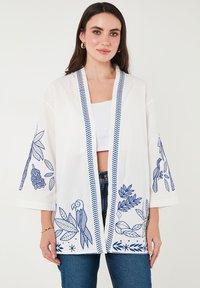Valge kimono jakk, millel on sinised brodeeritud lille- ja lindude mustrid, laiad varrukad ning mustriga viimistlus avatud ees.