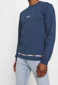 Námořnická modrá dlouhá rukávová tričko vyrobené z bavlny, s kulatým výstřihem, malou vyšitou nášivkou a potištěným názvem značky podél spodního lemu.