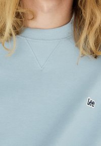 Sudadera azul clara con cuello de canalé, que presenta un pequeño logo bordado en navy de "Lee" en el lado izquierdo del pecho y un detalle triangular texturizado.