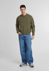 Pull en tricot olive avec un col en côtes, associé à un jean large bleu et des baskets grises. Design simple et texture douce.