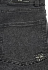 Svart denimtyg med en slät yta, som har en bakficka och en vävd etikett med en logotyp i vitt och svart.