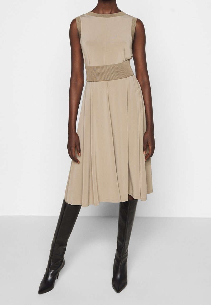 Une robe sans manches beige avec une taille côtelée ajustée, une jupe fluide et une texture lisse, associée à des bottes noires à talons pointus.