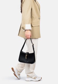 Borsa in pelle nera con parte superiore curvata, accessori in oro e una tracolla corta, tenuta da una persona in un blazer beige e pantaloni crema.