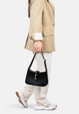 Sac en cuir noir avec un dessus arrondi, quincaillerie en or et une courte bandoulière, tenu par une personne portant un blazer beige et un pantalon crème.