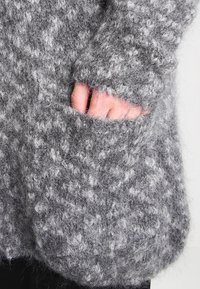 Šedý pletený svetr s nadýchanou texturou, který má přední kapsu a volný střih. Materiál vypadá měkký a teplý.