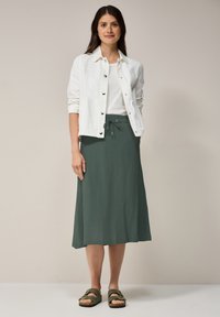 Witte overhemdjas boven een wit T-shirt, gecombineerd met een donker groene rok tot de knie en olijfgroene sandalen met gespen. Glad textuur.