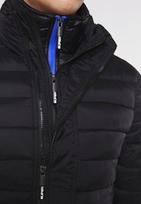 Sort puffjakke med høj krave, der har horisontale quiltede linjer, sorte lynlåse og en blå accentlynlås. Glat, syntetisk materiale.
