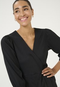 Černé šaty s nabíranými rukávy, hlubokým výstřihem do V, texturovanou látkou a přiléhavým pasem. Významné detaily zahrnují boční kapsy a jednoduchý design.