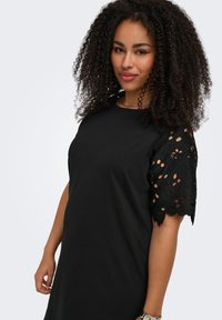 Woman with curly hair wearing a black dress with floral lace short sleeves, standing against a plain light background.