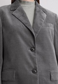 Gray corduroy blazer features a notched lapel, two buttons, and a chest pocket, paired with a light gray ribbed turtleneck underneath.