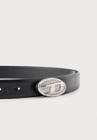 Melnas ādas josta ar ovālu sudraba sprādzi, kas izrotāta ar mazām kristālu akcentēm un stilizētu "D" dizainu uz baltas fona.