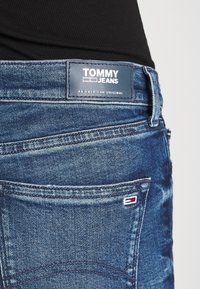 Vaqueros de mezclilla azul con un parche de cuero que presenta el logo "TOMMY JEANS". Costuras visibles, diseño del bolsillo trasero y pequeño acento de bandera cerca del bolsillo.