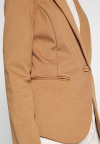 Gros plan d'une personne portant un blazer couleur chameau avec un bouton et une poche passepoilée par-dessus un haut blanc côtelé.