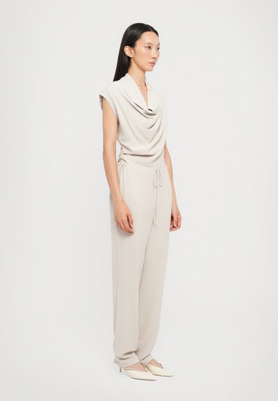 Cowlneck jumpsuit in lichtbeige stof, met korte mouwen, een trekkoord in de taille en taps toelopende benen, gecombineerd met spitse witte schoenen.