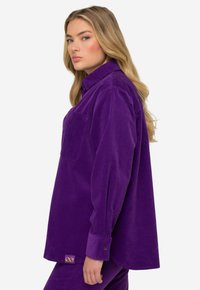 Violetti corduroy-paita, jossa on rintatasku, nappikiinnitys hihansuissa ja löysä leikkaus. Huomattava rakenne ja pehmeä pintakäsittely. Kullanvärinen logo yksityiskohtana helmoissa.