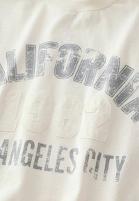 Světlobéžová mikina s opotřebovaným šedým potiskem s nápisy "CALIFORNIA 1982" a "LOS ANGELES CITY" v různých velikostech písma.
