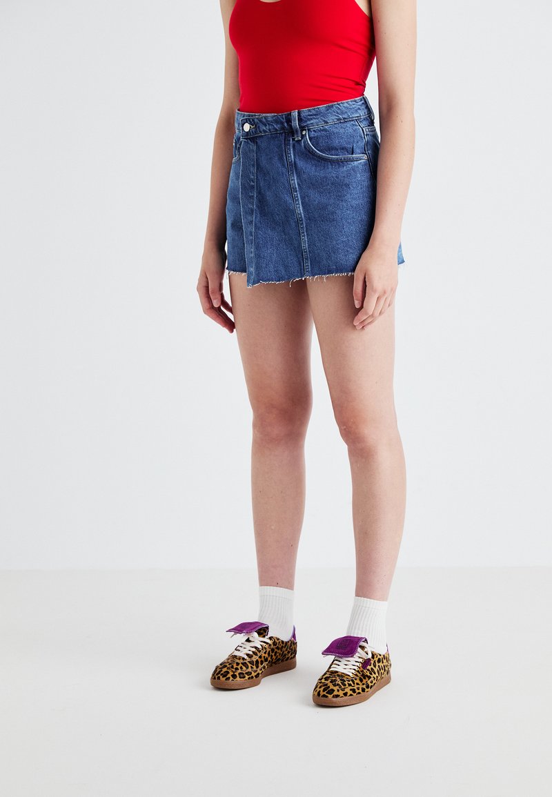 Denim mini szoknya nyers szegéllyel, világoskék színben, leopárdmintás cipőkkel, amelyeken lila díszítések vannak, fehér zoknival párosítva.