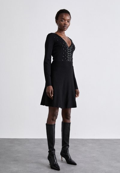 Vestito nero con maniche lunghe, scollatura a V profonda, impreziosito con borchie sul busto. Gonna a tulipano svasata e vita aderente. Stivali con tacco fino al ginocchio.