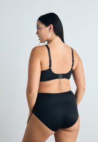 Soutien-gorge noir et culotte taille haute présentés de dos, avec des bretelles réglables et une fermeture à crochets. Texture de tissu lisse.