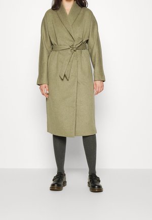 Femme portant un manteau ceinturé olive longueur genou, des leggings gris foncé et des chaussures noires à boucle, debout sur un fond blanc uni.
