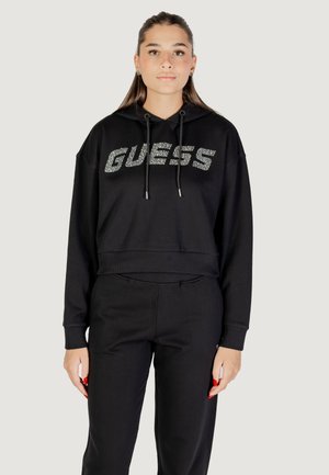 Hoodie corto nero con la scritta "GUESS" in strass, con cordini e polsini a costine. Abbinato a pantaloni neri coordinati.