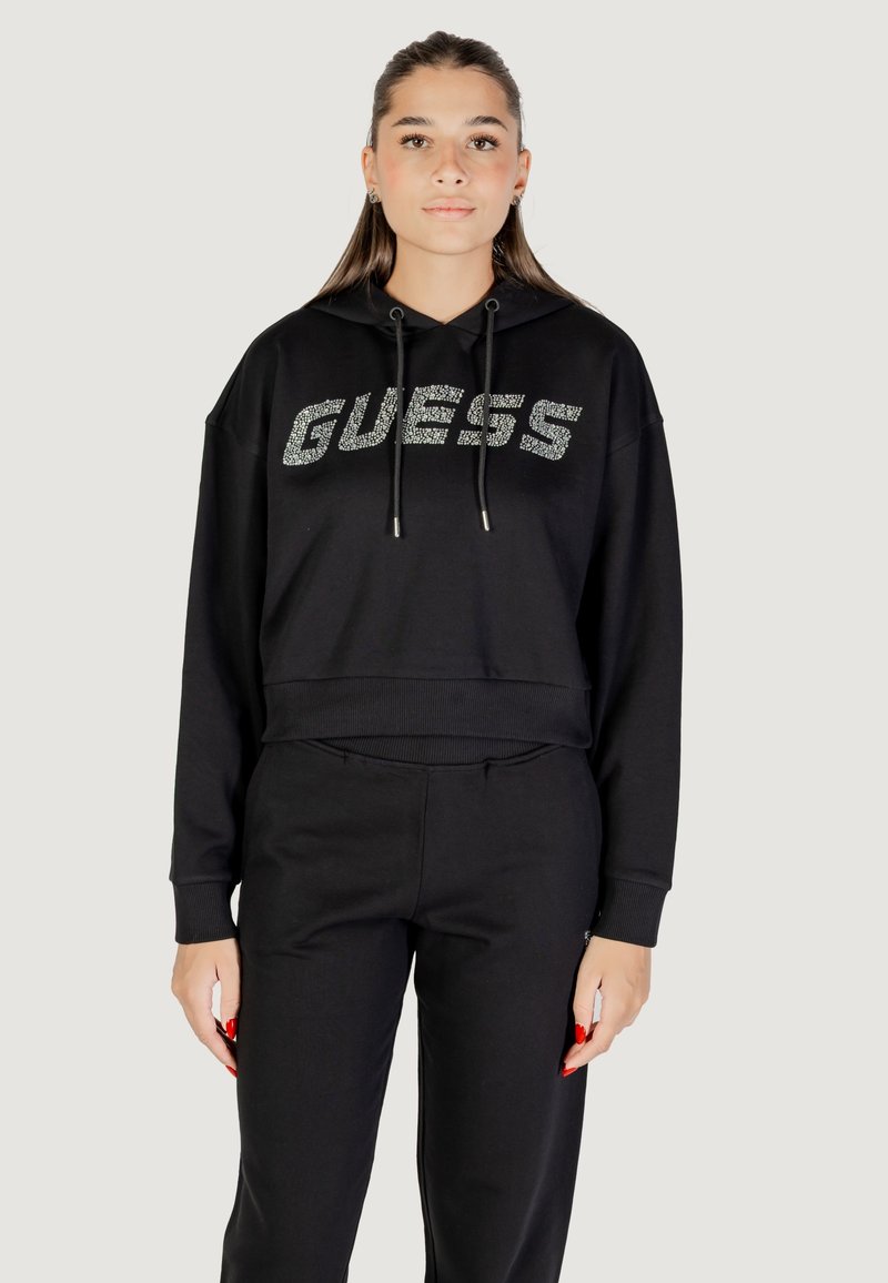 Haut de survêtement cropped noir avec le mot "GUESS" en strass, doté de cordons de serrage et de poignets côtelés. Associé à un pantalon noir assorti.