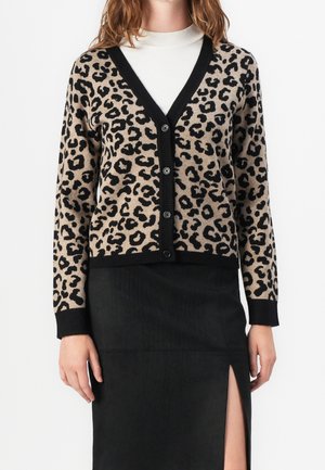 Femme portant un cardigan beige et noir à imprimé léopard boutonné sur un top blanc, associé à une jupe noire fendue devant.