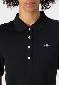 Polo negru cu guler, având o clapetă cu butoane albe și un mic logo brodat pe pieptul stâng. Material texturat.